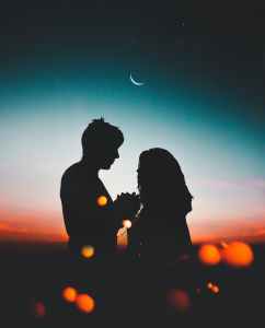 Silhouette of two people holding hands, crescent moon in evening sky above.