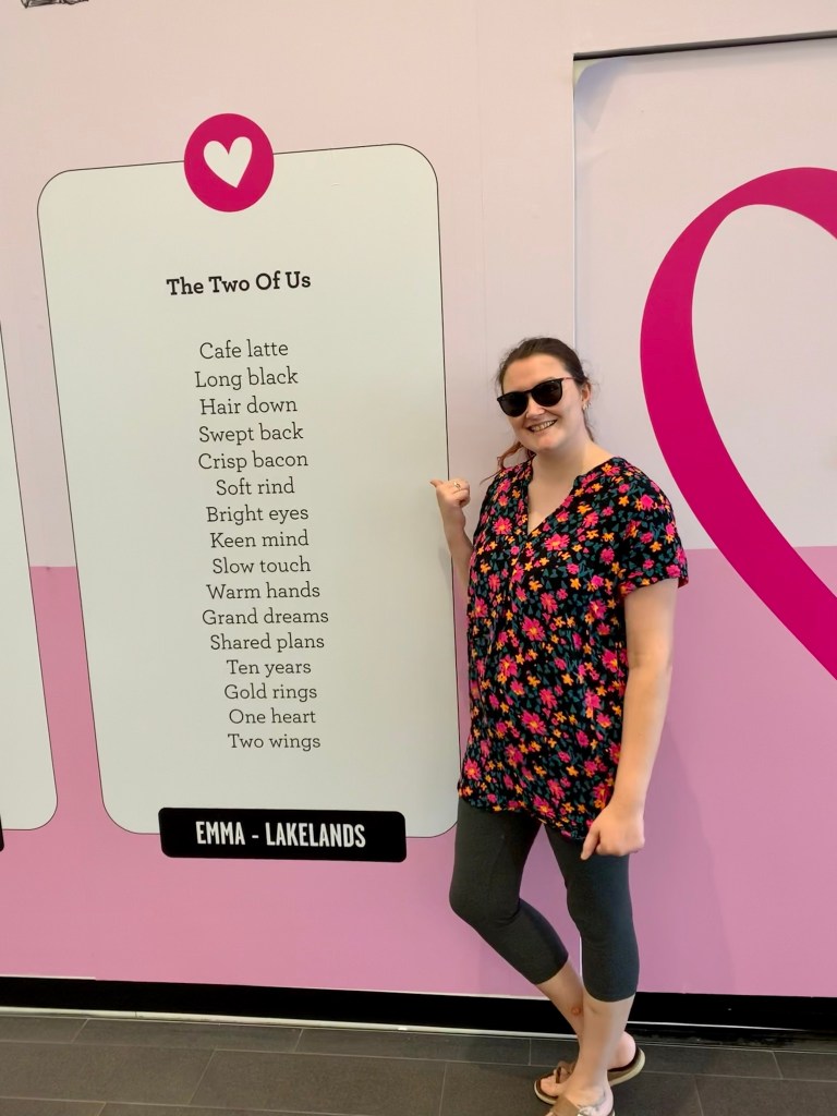 Woman in sunglasses, leggings and bright top pointing to a full-length wall poster of a poem.