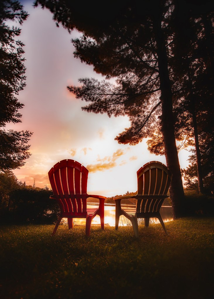 Two adirondack style chairs on grass, facing away towards a body of water, trees nearby, at sunset