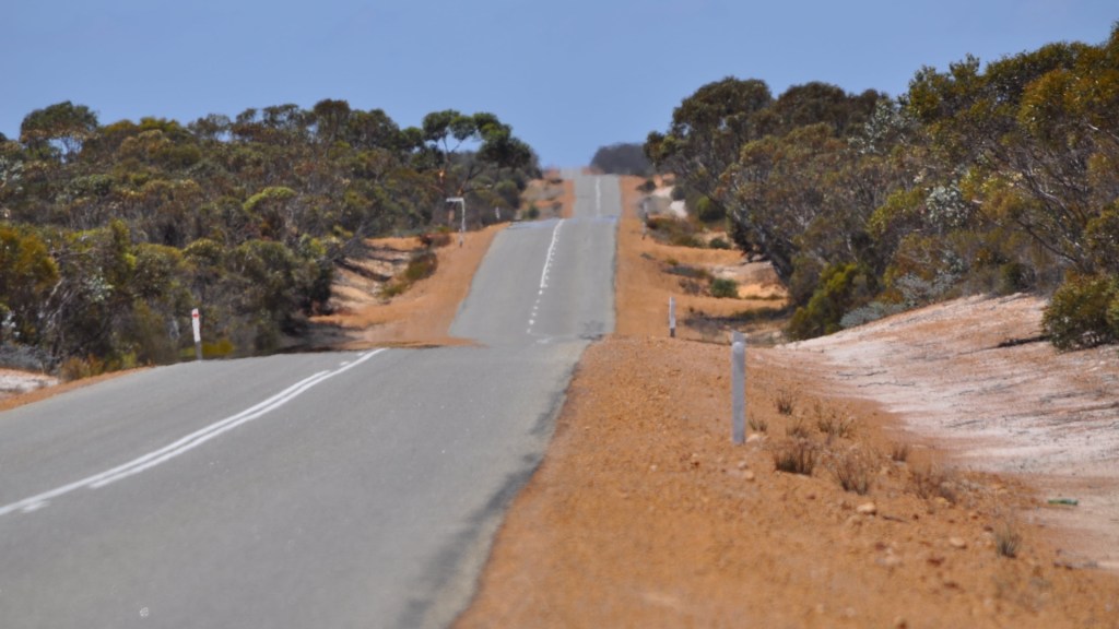 Asphalt road with white dividing line travelling from near to far distance over small hills, red and white dirt to each side, green Australian bush plants, pale blue sky