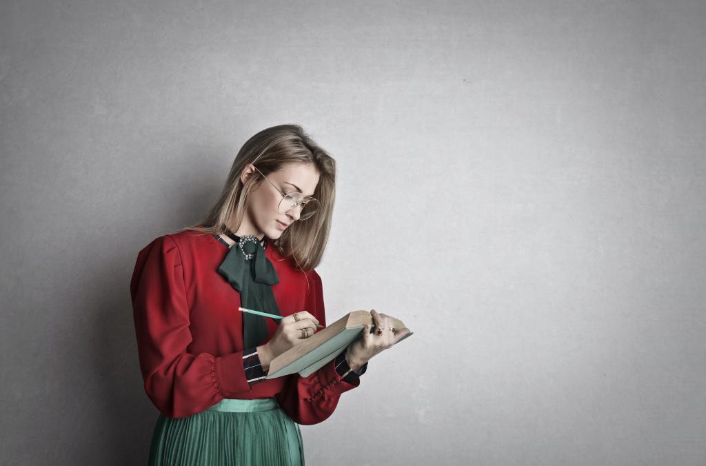 Smart adult female student reading a book while standing, holding a pencil in one hand poised to write on the page.