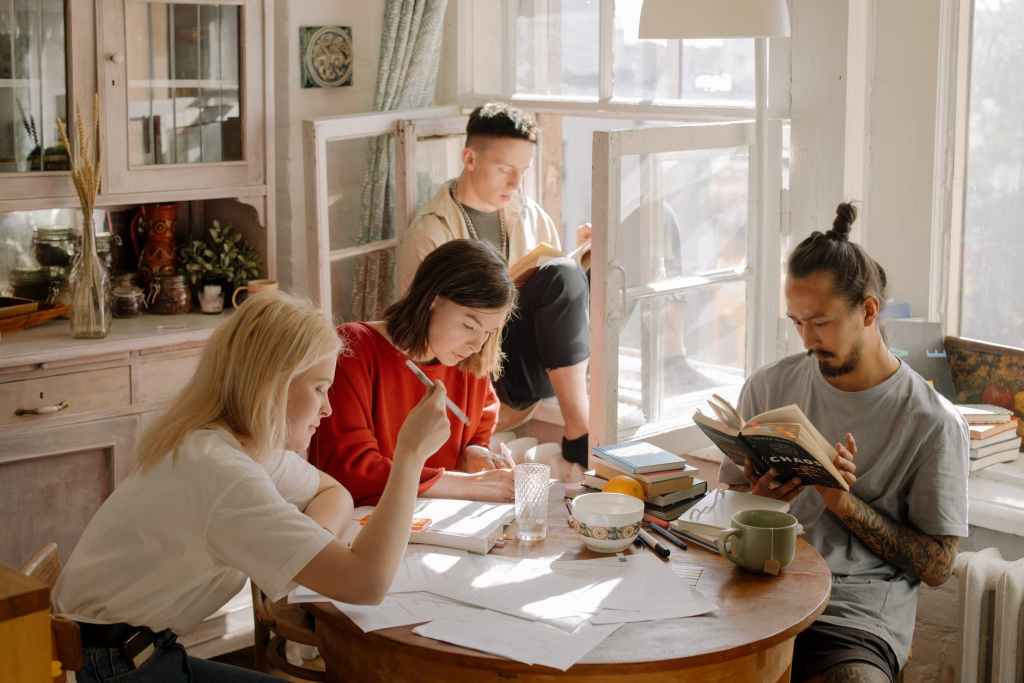 group of four young adults in a kitchen with various books, paper, texts, writing implements and refreshments scattered about