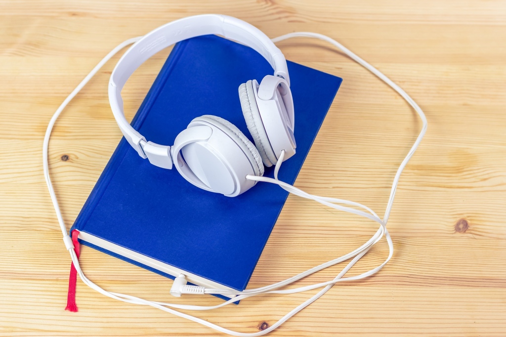 Book with blue cover, closed, on a pale wood surface, with white large headphones and cord on top of the book 
