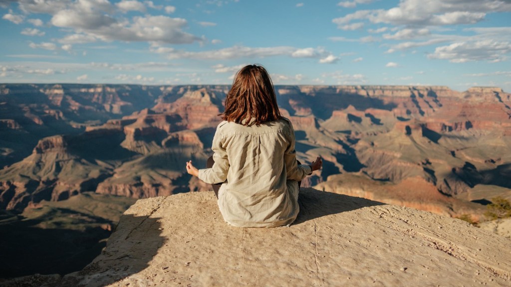 Person seated on a ledge, shirted back to photographer, legs crossed arms on legs in meditation pose, shoulder length brown hair down, looking out over a view of a canyon system.