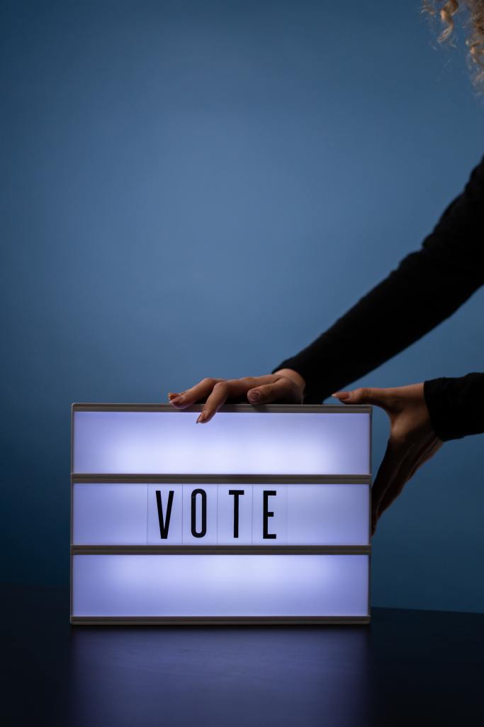 white sign with VOTE written on it, lit from within (lightbox), with hands holding it on a table - a pair from someone with white skin and red painted nails