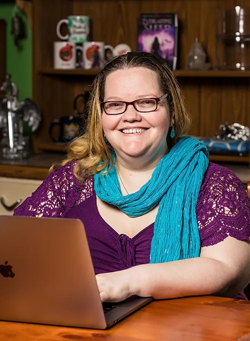 Image of author Rebecca Laffar-Smith, a smiling white woman with glasses, blonde-brown hair worn down, a green scarf and red top with an open laptop in front of her on a desk