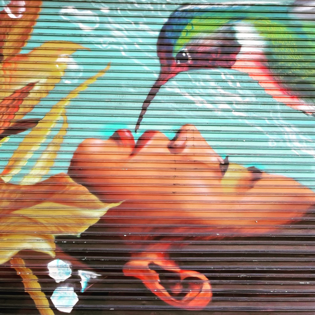 Image of a painted mural on a garage door of a prone, medium-skinned woman's face receiving dew from a larger-than-life hummingbird